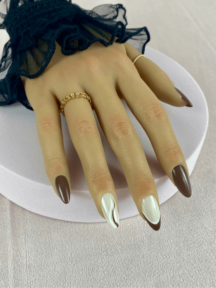 Mains portant des faux ongles réutilisables adhésifs de couleurs marron avec un premier nail art qui a deux lignes, une blanche et une marron puis le deuxième nail art à une French manucure marron avec une ligne blanche. 